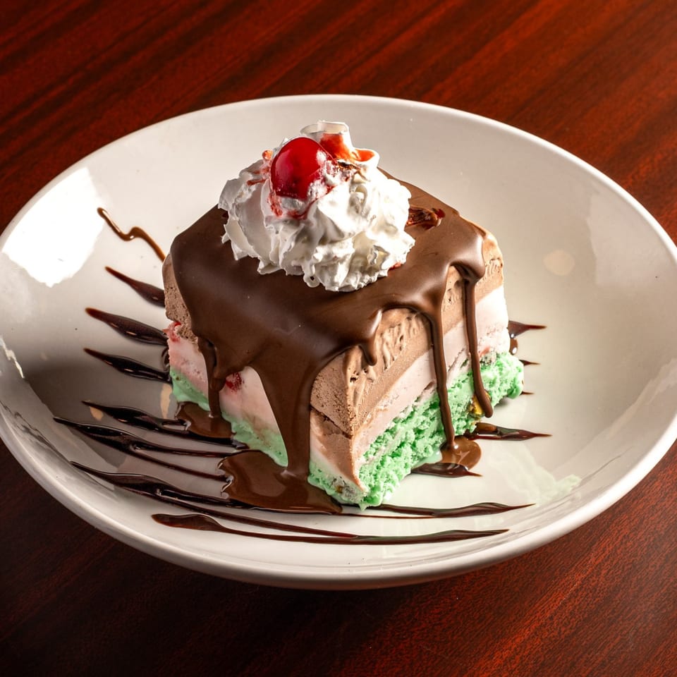 A slice of layered spumoni ice cream with chocolate sauce, whipped cream, and a cherry.