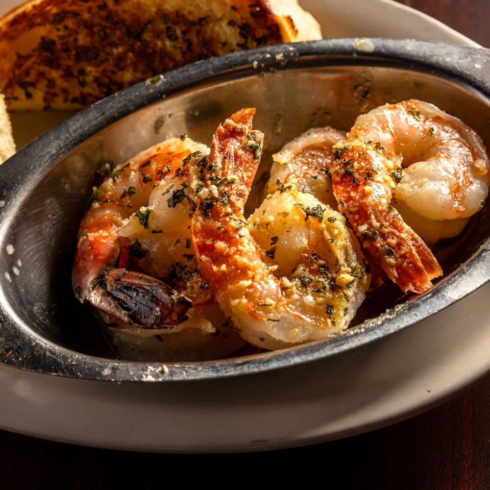 Garlic shrimp appetizer with herbs in a metal dish, with toasted bread in the background.