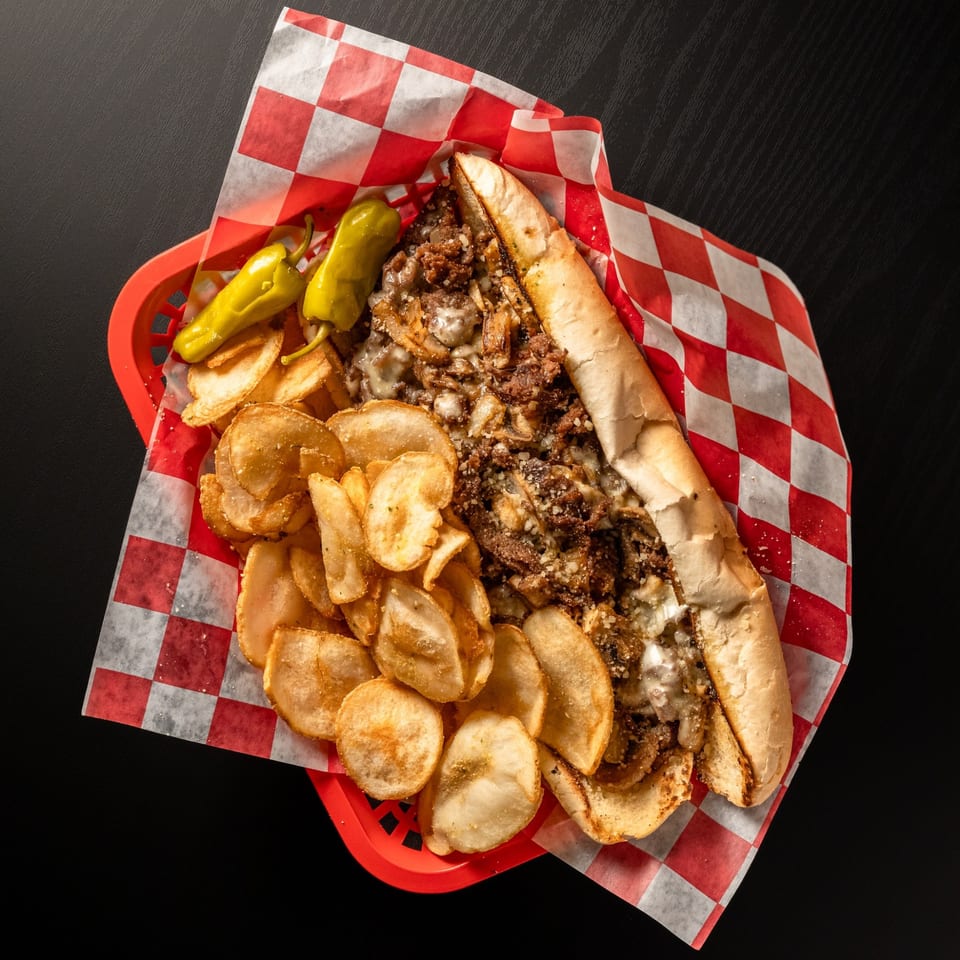Cheesesteak sandwich with chips and peppers in a red basket with checkered paper.